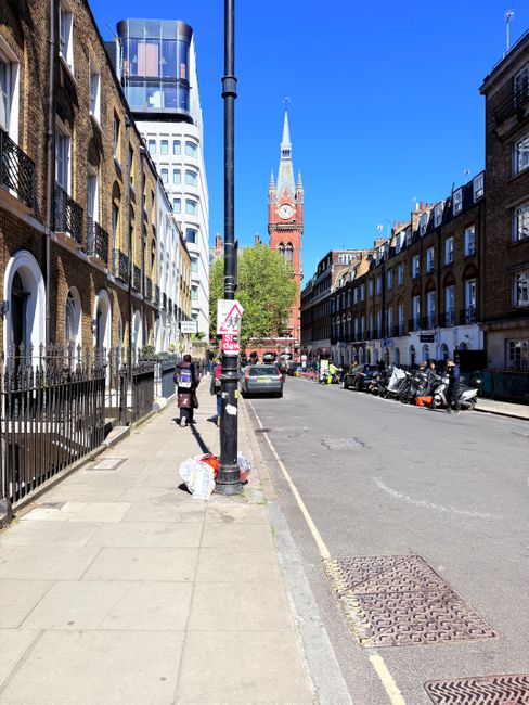 A wide view of a street in Bermondsey with residential buildings on both sides, featuring brick facades and multiple windows. The sidewalk on the left is paved with concrete slabs and has a metal fence bordering the building. A lamppost stands near the curb, with a plastic bag and a small garbage bin at its base. Several pedestrians are walking along the sidewalk, some carrying bags. On the right side, at least five parked motorcycles and a car are visible along the street. In the background, a prominent red brick clock tower with a pointed spire is set against a clear blue sky, with modern glass-fronted buildings nearby. The scene suggests a typical urban environment suitable for home relocation activities, where furniture and boxes may be loaded onto vehicles by professional removals companies like Bermondsey Removals, who handle packing and moving logistics around such street environments.