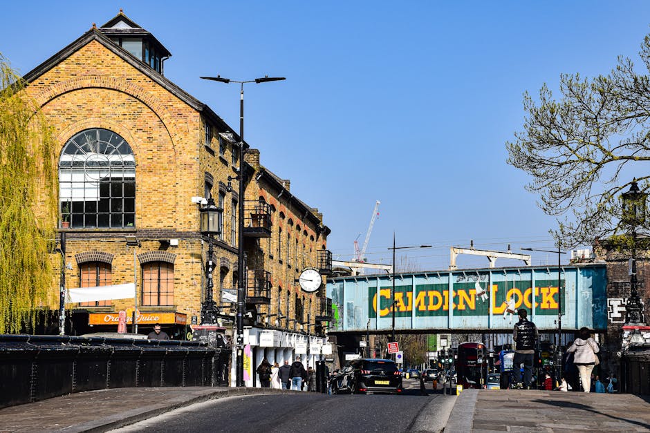 A street view in Bermondsey showing a brick building with large arched windows on the left side, adjacent to a pedestrian walkway with people walking. In the foreground, there are black metal railings and a sidewalk. A large, blue and yellow painted metal railway bridge with the words 'Calden Lock' spans across the street. Several black cars and a red double-decker bus are visible on the road, with some street lamps and leafless trees lining the street. The sky is clear and blue, and a construction crane can be seen in the background. The scene captures urban activity typical of a home relocation or furniture transport process, with possible loading areas nearby, and the scene is part of a Bermondsey Removals guide access tips page, illustrating transport and logistics in a city environment.