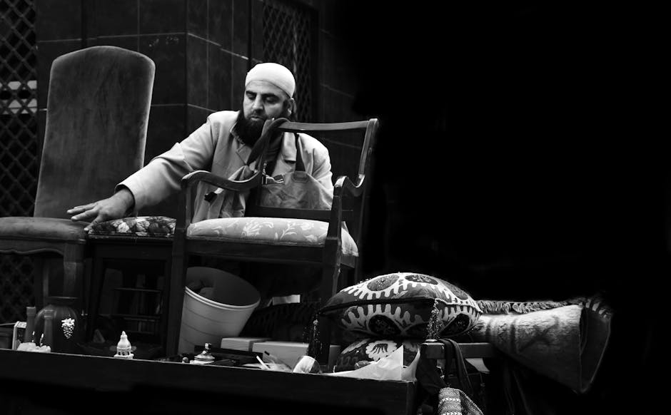 A man wearing traditional attire and a white head covering is performing a home relocation task, carefully lifting or adjusting a wooden chair with fabric upholstery, part of a collection of furniture including upholstered chairs, cushions, and a rolled-up carpet displayed on a flat surface. The background features a dark, textured panel, indicating an indoor setting, possibly a storage or staging area for moving items. Various packing materials such as cardboard boxes, plastic containers, and fabric wraps are visible around the furniture, suggesting packing and moving preparations. The scene is illuminated evenly, highlighting the detailed textures of the furniture and packing supplies. This image reflects the logistics involved in furniture transport and the organizational aspects of house removals, aligning with services offered by Bermondsey Removals for house and furniture movers advice, particularly for stalls at Maltby Street Market.
