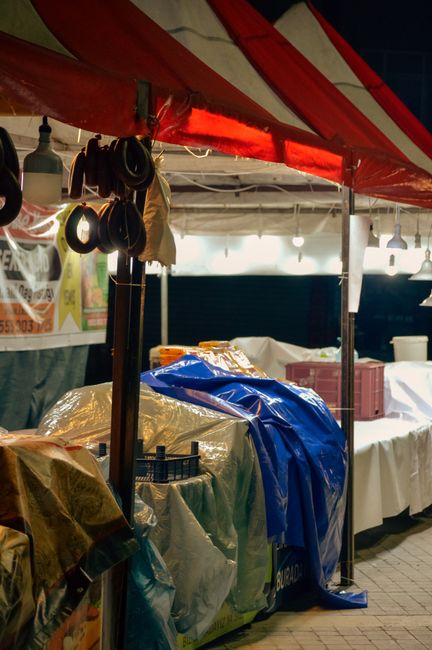 A covered outdoor market stall at night with a red and white striped canopy overhead. The stall features various items covered with plastic sheets, including large blue and transparent plastic coverings suggesting packed furniture or goods ready for transport. Hanging hooks with coiled black cables and a beige fabric item are visible from the canopy support pole. Behind the stall, there are tables covered with white cloths, some with cardboard boxes and other packaging materials, indicating packing and moving preparations. The scene is illuminated by hanging lightbulbs providing even lighting, highlighting the organized packing process typical of furniture transport or house removal setups. The environment appears to be part of a street market, with the presence of packaging materials and protective coverings aligning with professional removals or residential relocation activities, such as those coordinated by Bermondsey Removals in support of house moves or furniture transport at Maltby Street Market.