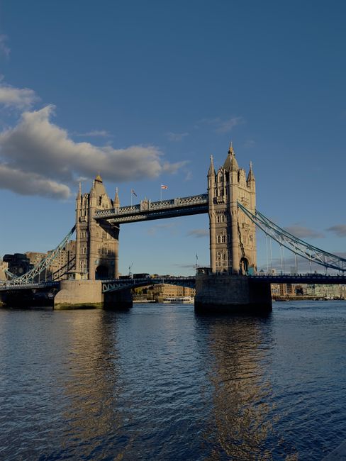 A view of Tower Bridge spanning the River Thames in London, with its iconic twin towers and blue suspension elements prominently visible. Several boats and ferries are seen navigating the river below,